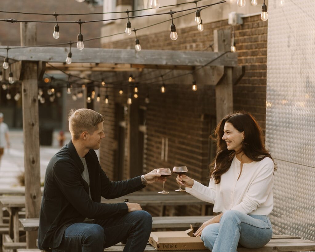 Engaged couple sitting on top of a picnic table in the city and doing a cheers with their glasses of wine