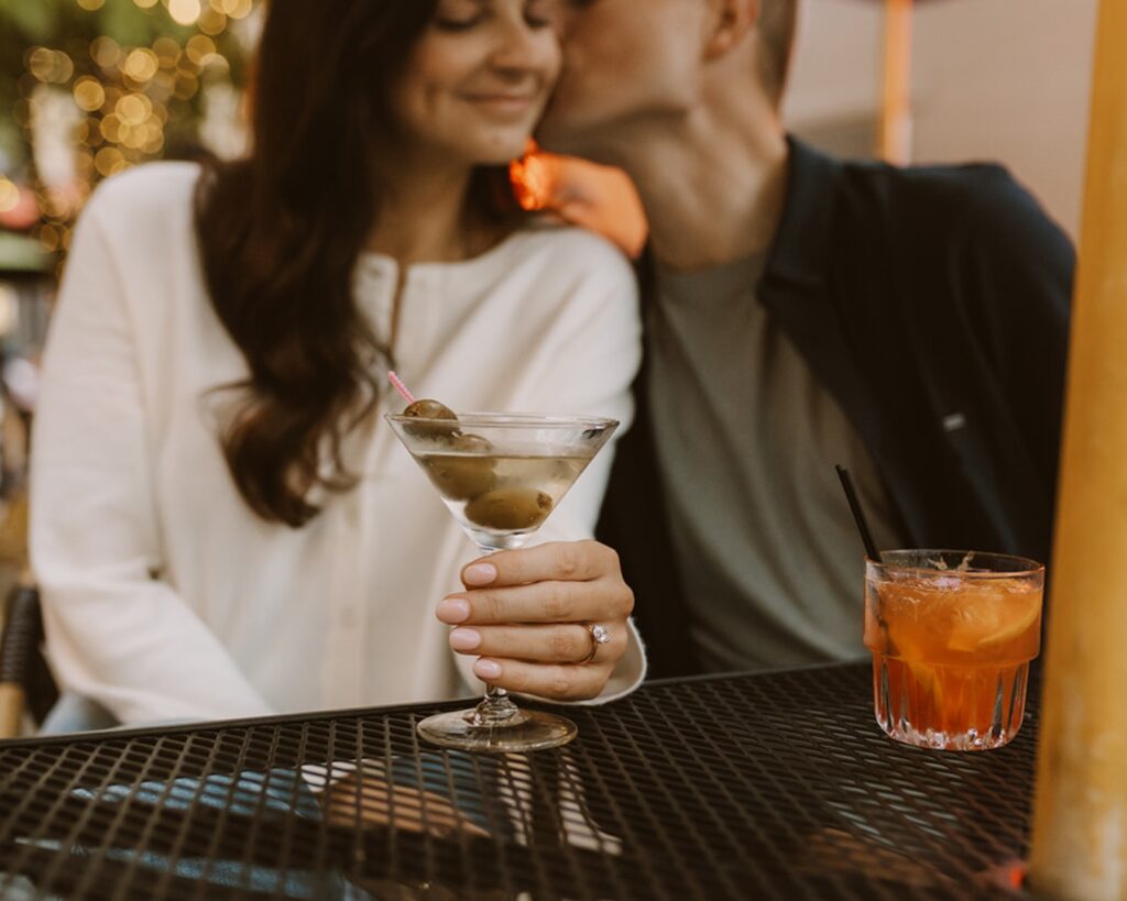 close up image of the engaged couples drinks as the groom to be kisses the bride to be on the cheek in the background