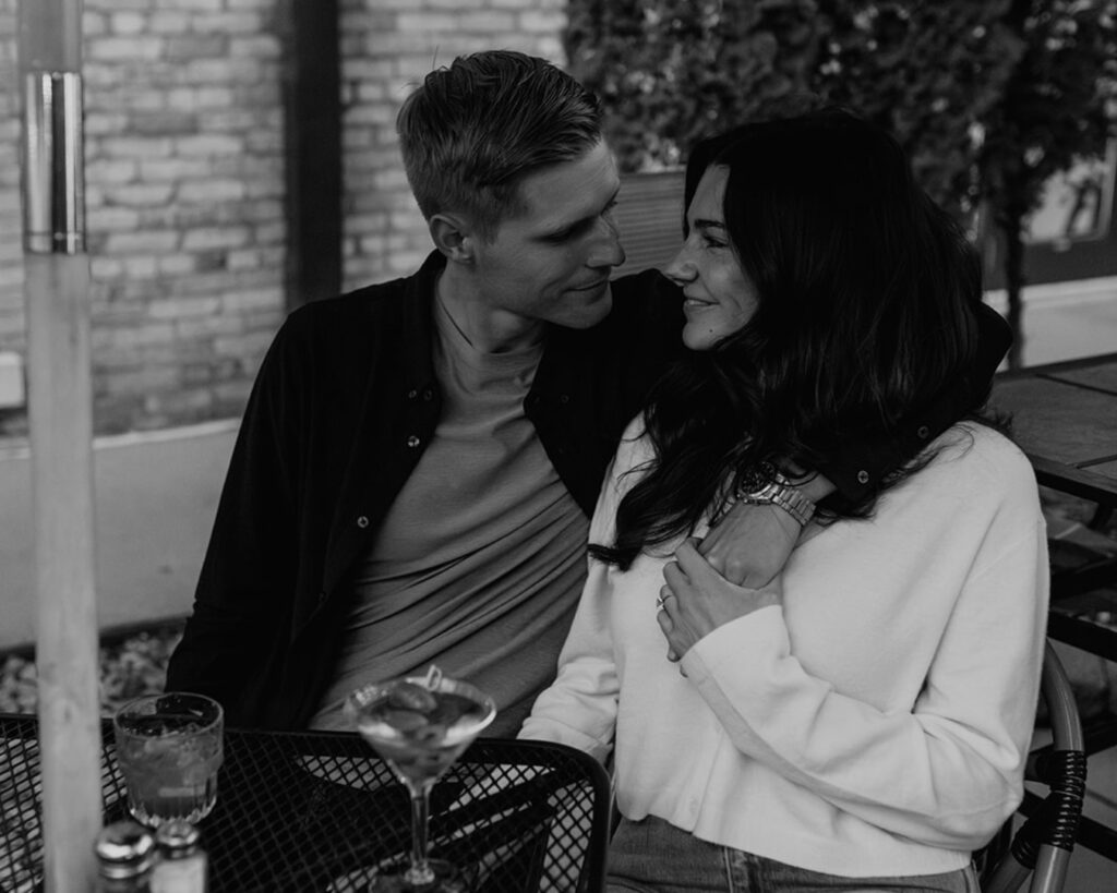 black and white image of an engaged couple snuggled up at a table on the patio of their favorite restaurant as they share a drink