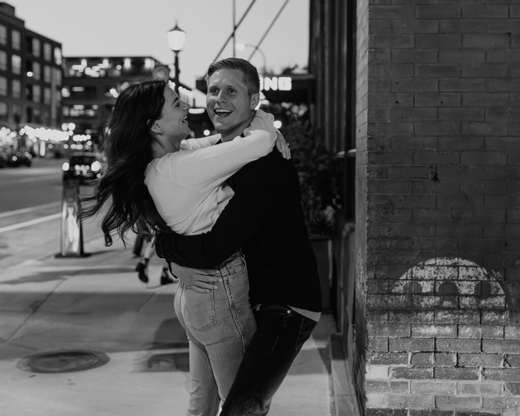 Black and white image of an engaged couple embracing and laughing on a city street corner 