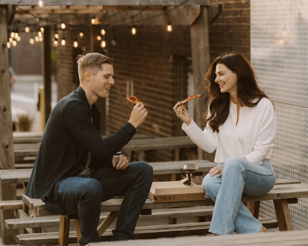 Engaged couple sitting on top of a picnic table in the city while they eat pizza and chat