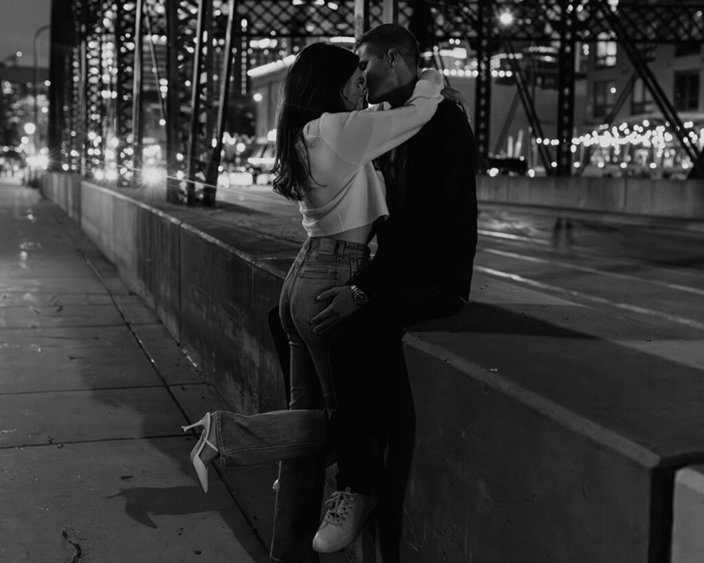 black and white image of an engaged couple snuggling up  on a bridge with the city lights behind them