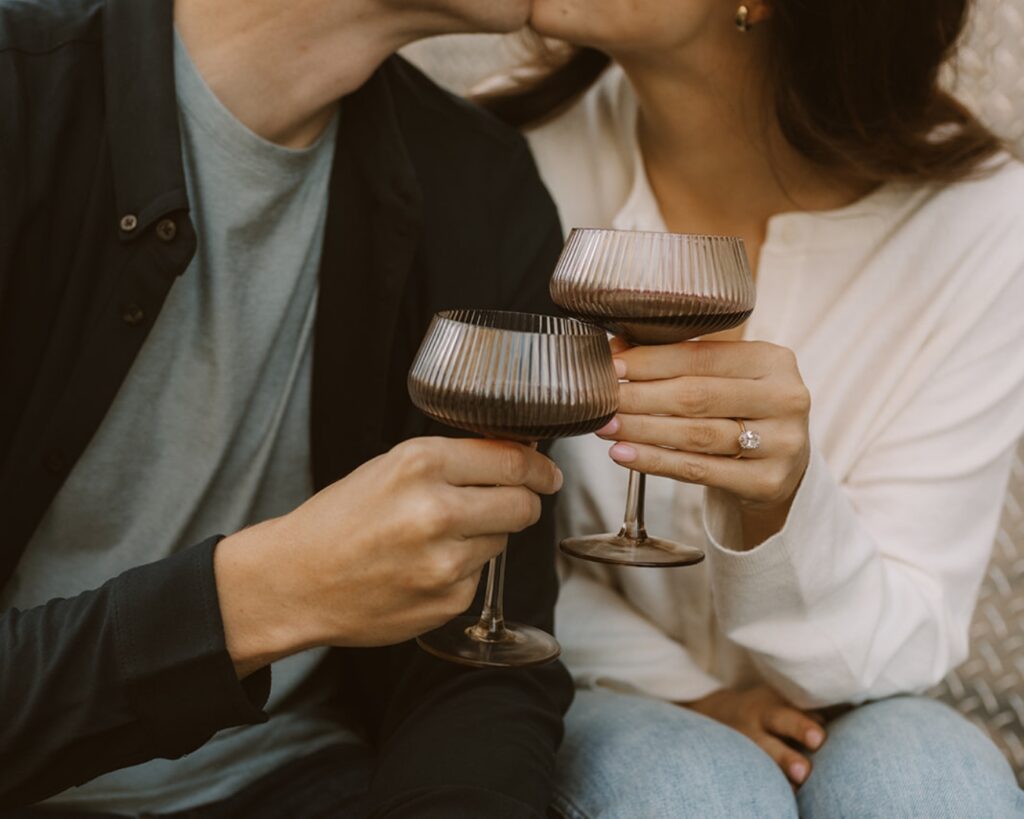 close up image of the engaged couple's hands as they clink their glasses of wine together to show off the bride to be's engagement ring 