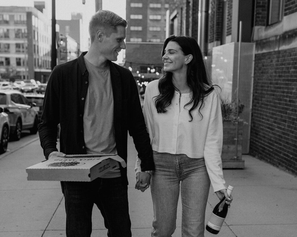 black and white image of an engaged couple walking in the city holding a boxed pizza and a bottle of wine 
