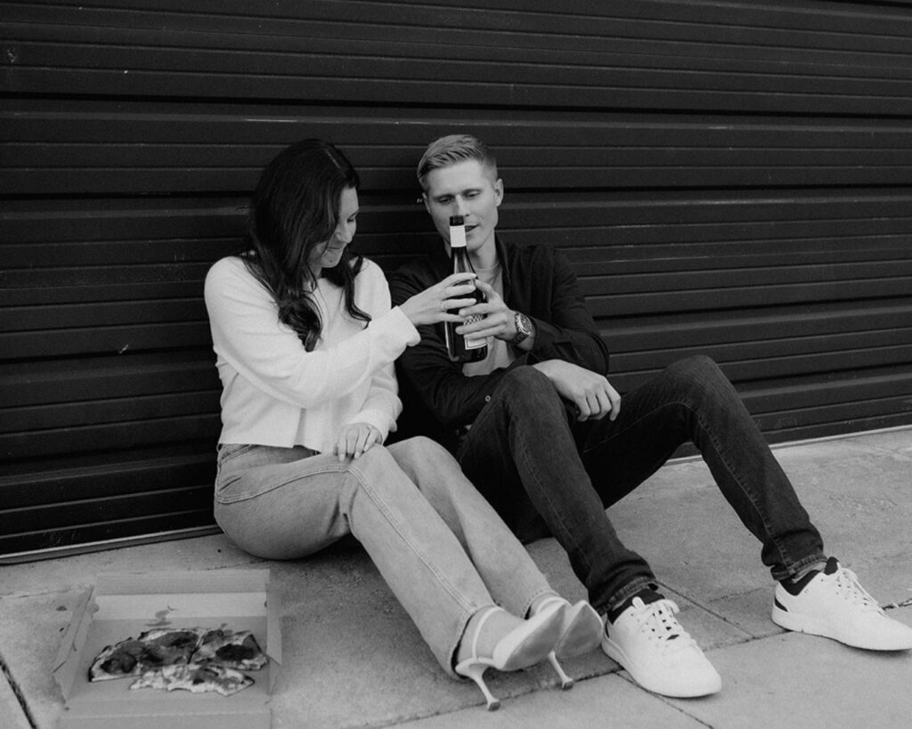 Engaged couple sitting on the ground against a building and sharing a bottle of wine and a pizza