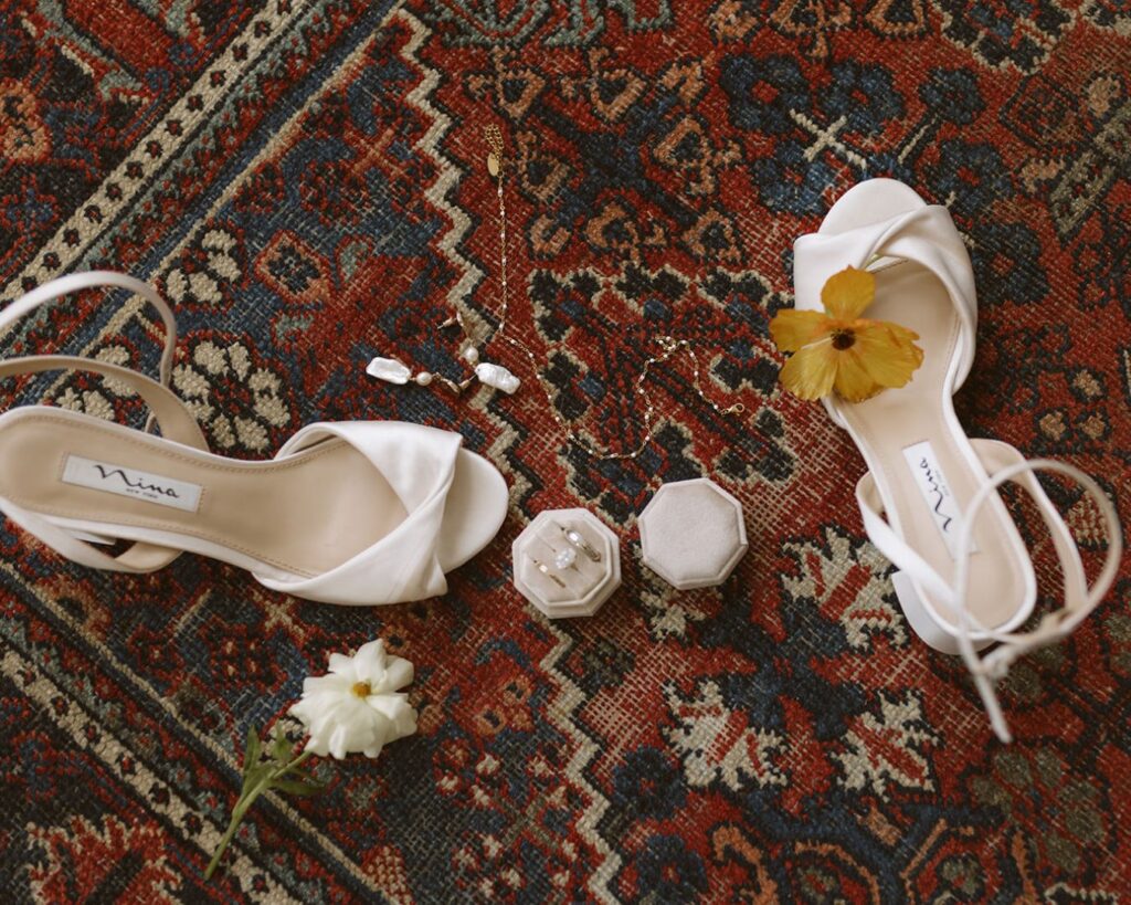 The bride's details laid out on an ornate rug. Details including shoes, a flower, the rings in a ring champagne ring box, her earrings, and necklace