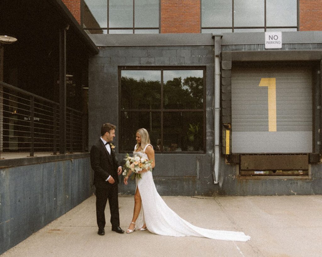 the bride stepping towards the groom as she holds her bouquet in an industrial area outside their venue 