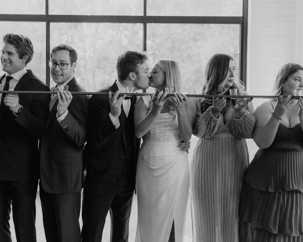 Black and white image of the bride and groom kissing while lined up at shotski with some of their friends