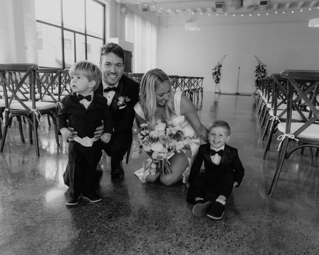 the bride and groom kneeling down on the floor to take a photo with their two ring bearers. The bride is smiling at the littlest ring bearer who is sitting on the ground smiling right at the camera 