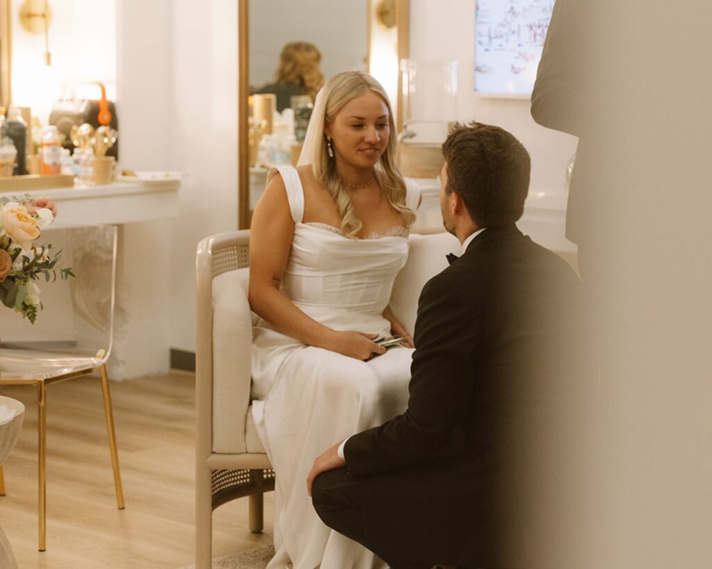 the bride is sitting in a chair in the bridal suite with the groom knelt in front of her as they have a moment just before walking down the aisle