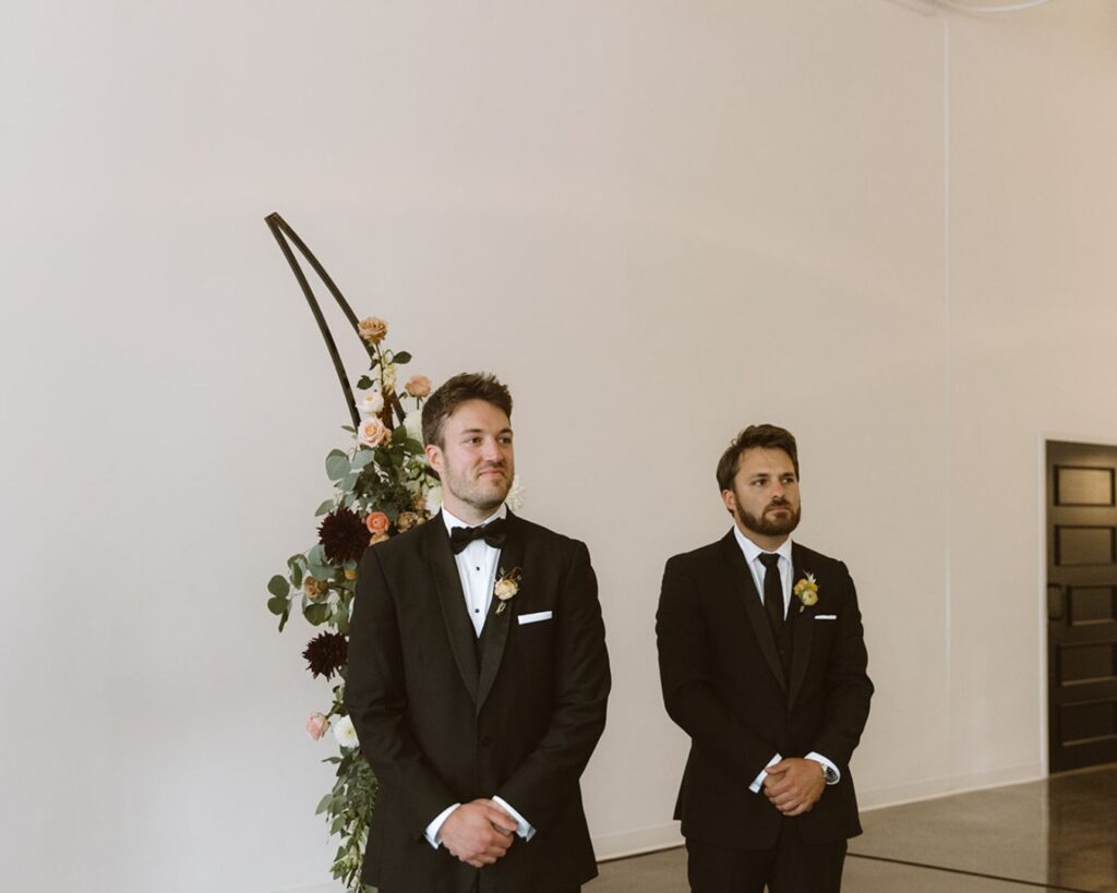 The groom smiling as he watches his bride make her way down the aisle with her parents on each arm