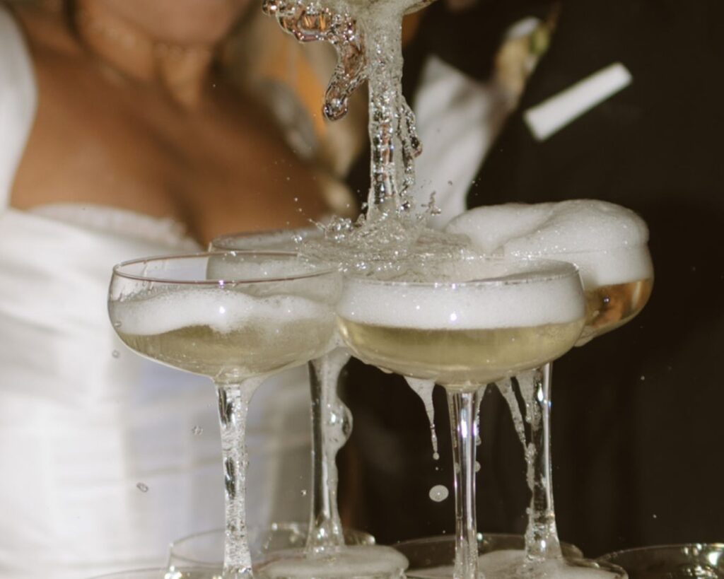 Up close shot of champagne hitting the top glasses of the champagne tower and splashing all over. You can see just a bit of the bride and groom in the background as they pour. 