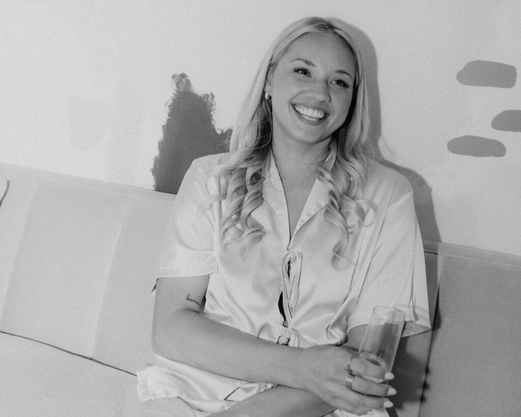 Black and white image of the bride smiling while sipping champagne on the couch in her getting ready outfit