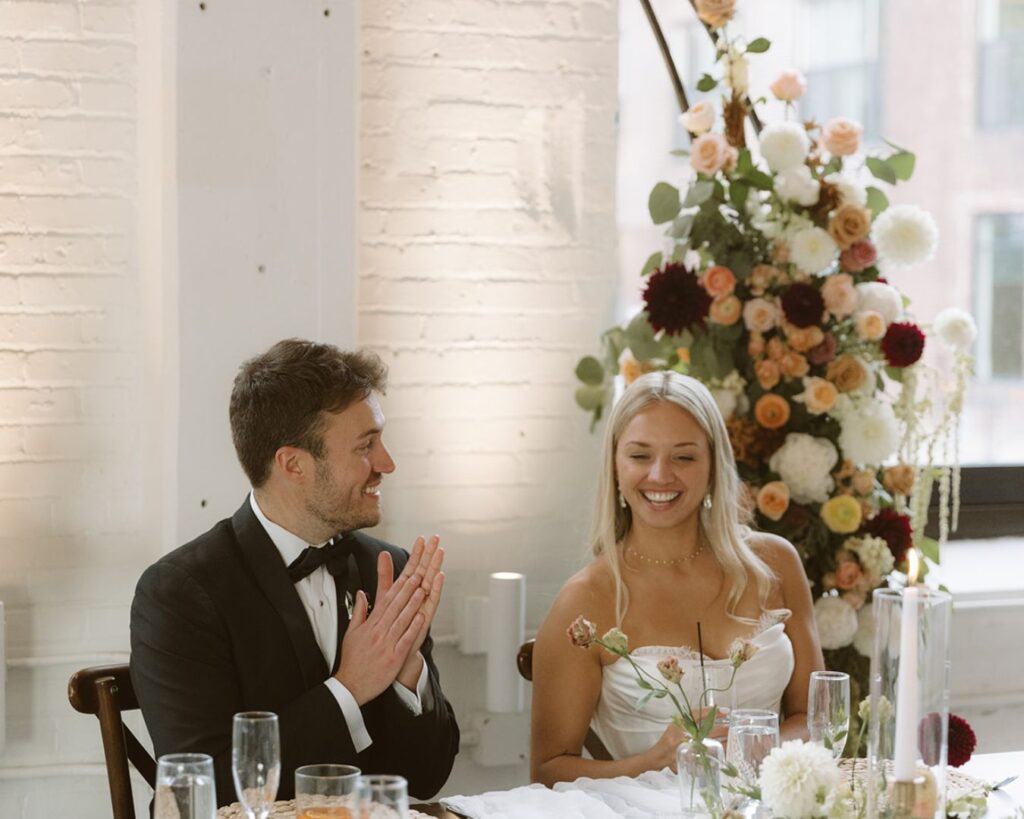 The bride and groom sitting at their head table after viewing their reception space before anyone has entered it. They are talking and smiling. 