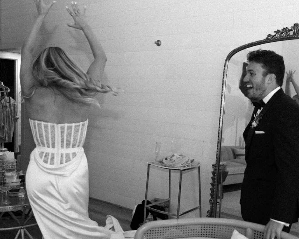 a black and white image of the bride twirling in the bridal suite as her groom watches her with a smile on his face