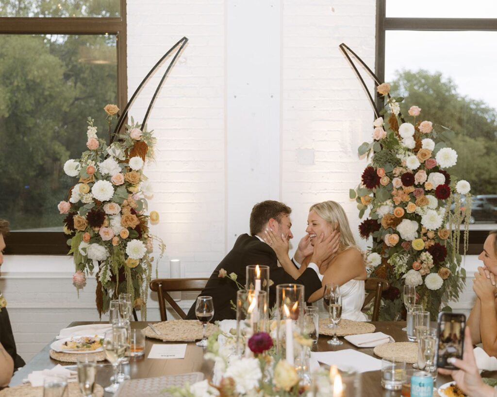 The bride and groom about to kiss as they sit at their head table during the reception. They are both grabbing onto each others faces with both hands. 