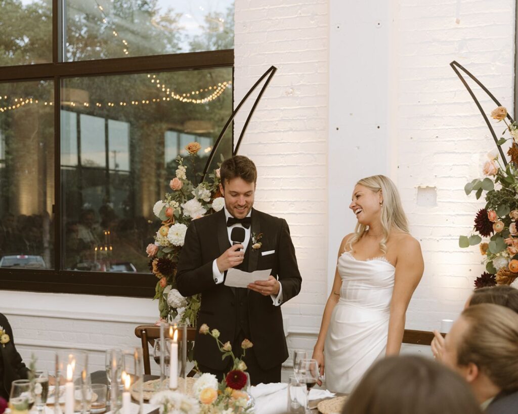 The bride smiling at the groom as he reads his speech. 