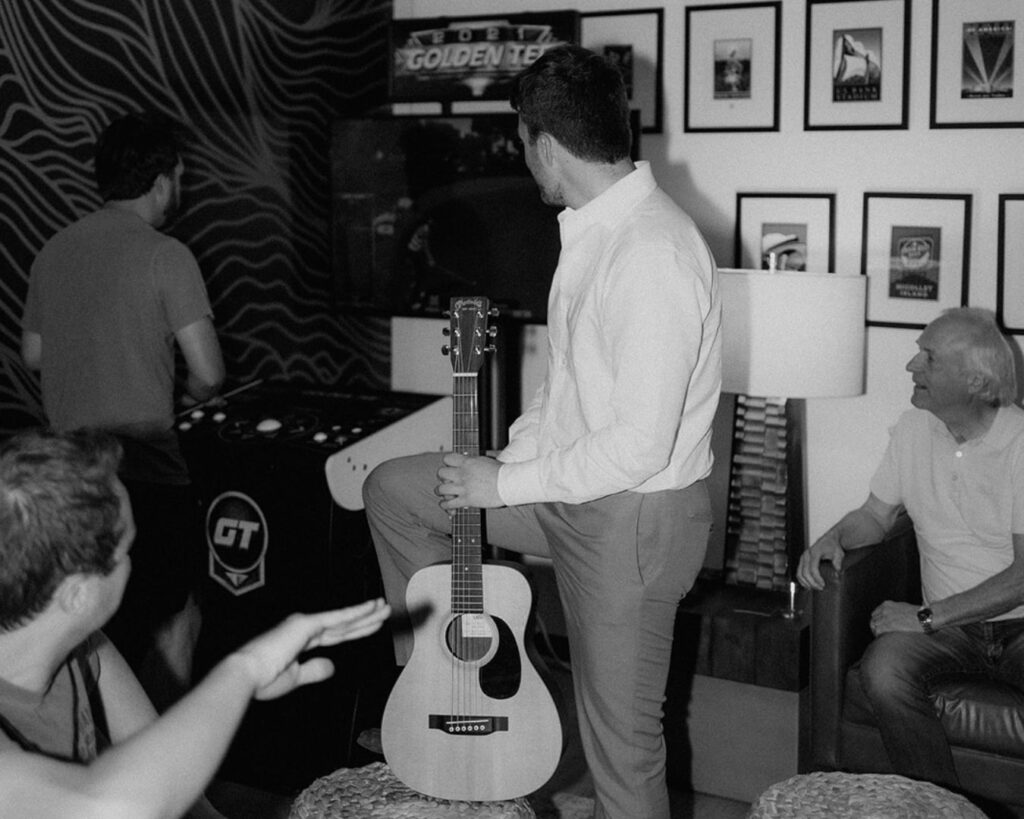 Black and white image of the groom holding a guitar while watching a friend play an arcade game