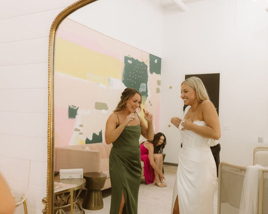 the bride and her maid of honor giggling as she puts on her dress