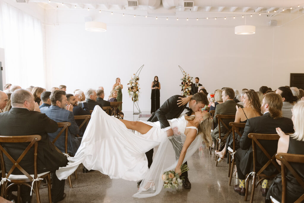 The bride and groom kissing in the middle of the aisle just after being announced as husband and wife. Their guests are cheering and clapping all around them 