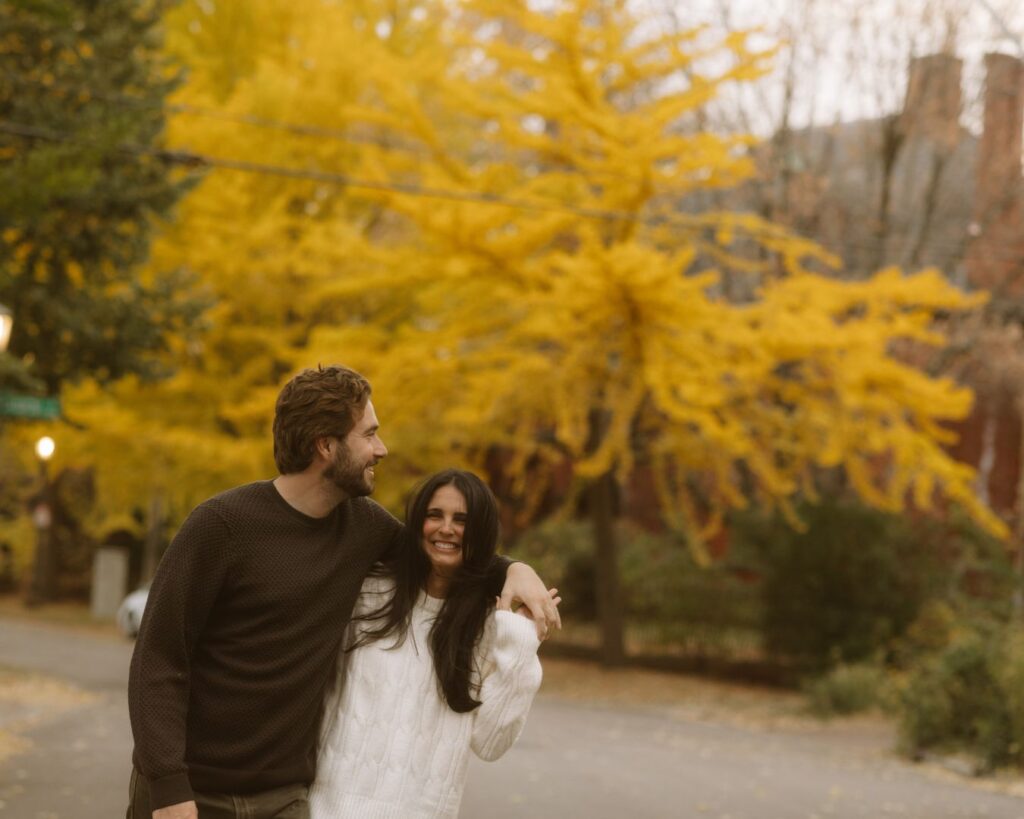 Couple laughing and walking arm-in-arm in Cathedral Hill, St. Paul, with vibrant yellow autumn leaves and historic buildings in the background.