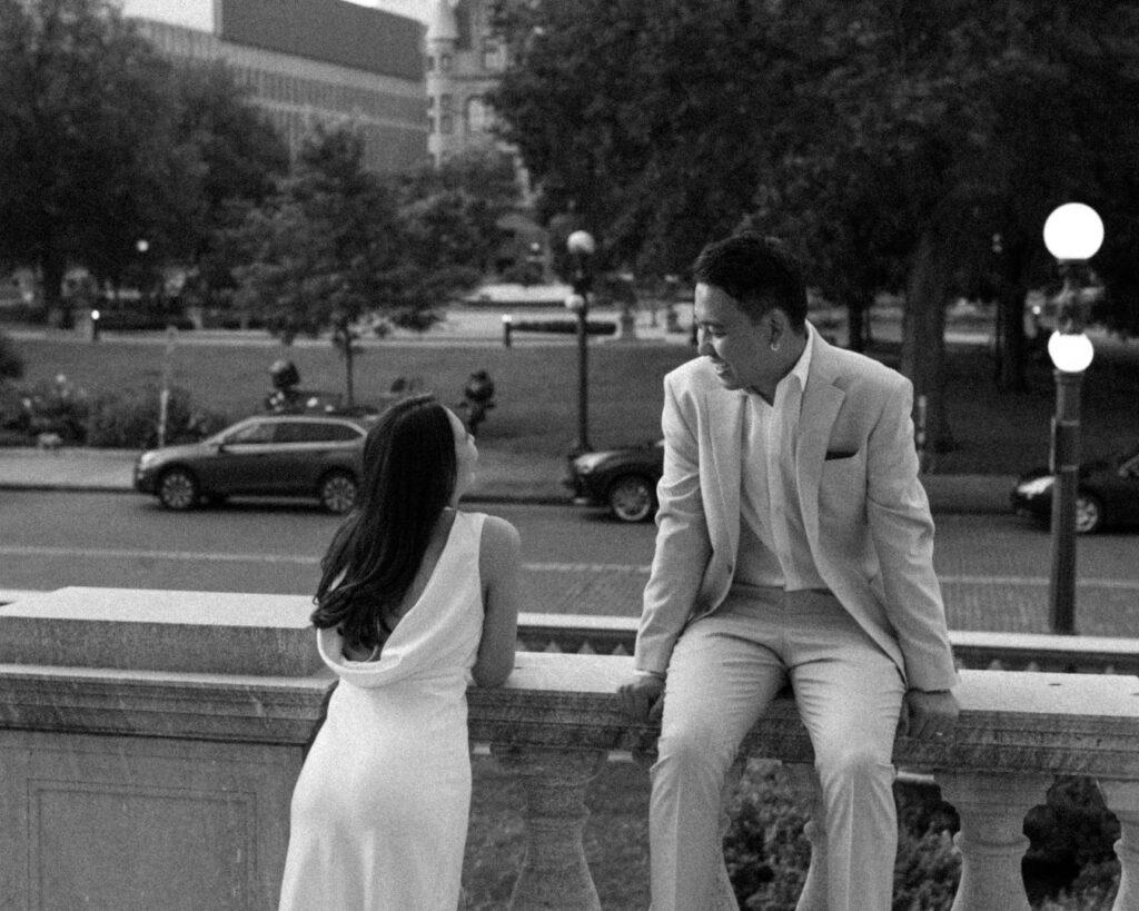 Black and white image of a couple chatting while he sits on a ledge and she leans up against it in the city 