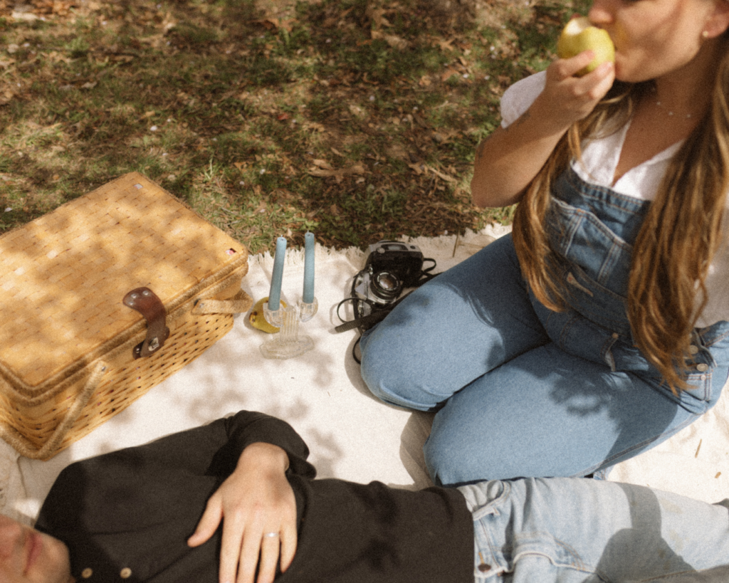 Couple on a picnic blanket. He is laying down and she is sitting on her knees eating a pear. 