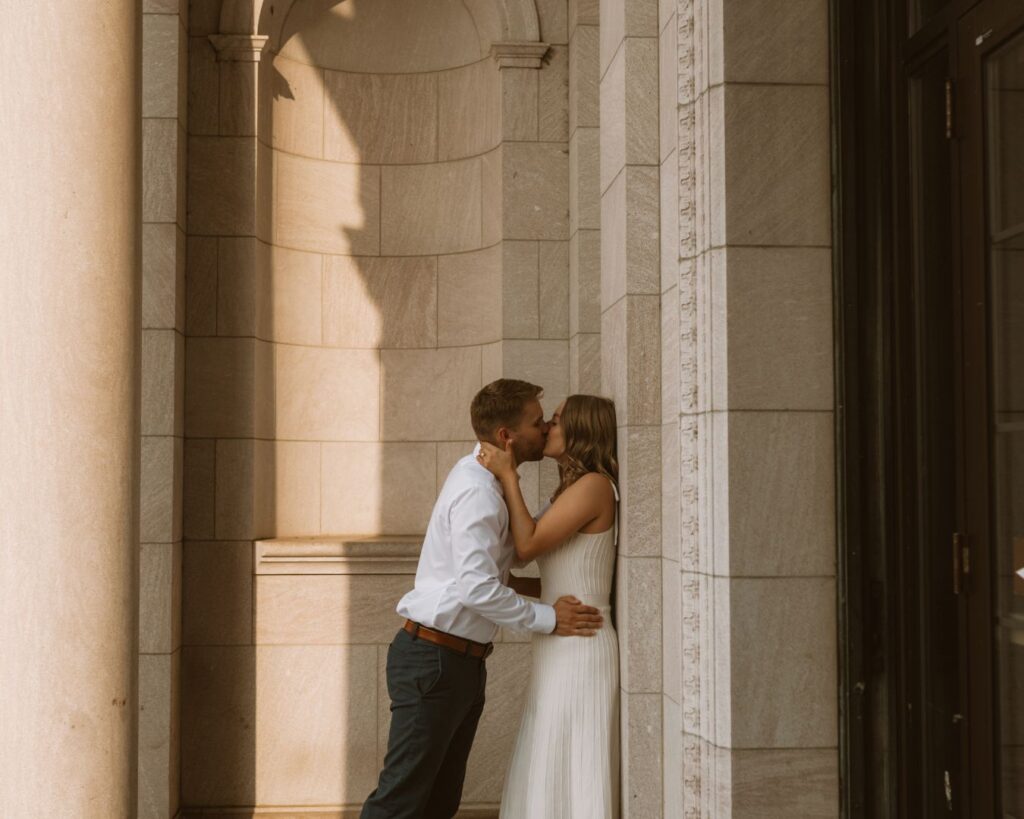 A couple leaning up against a light stone wall kissing. 