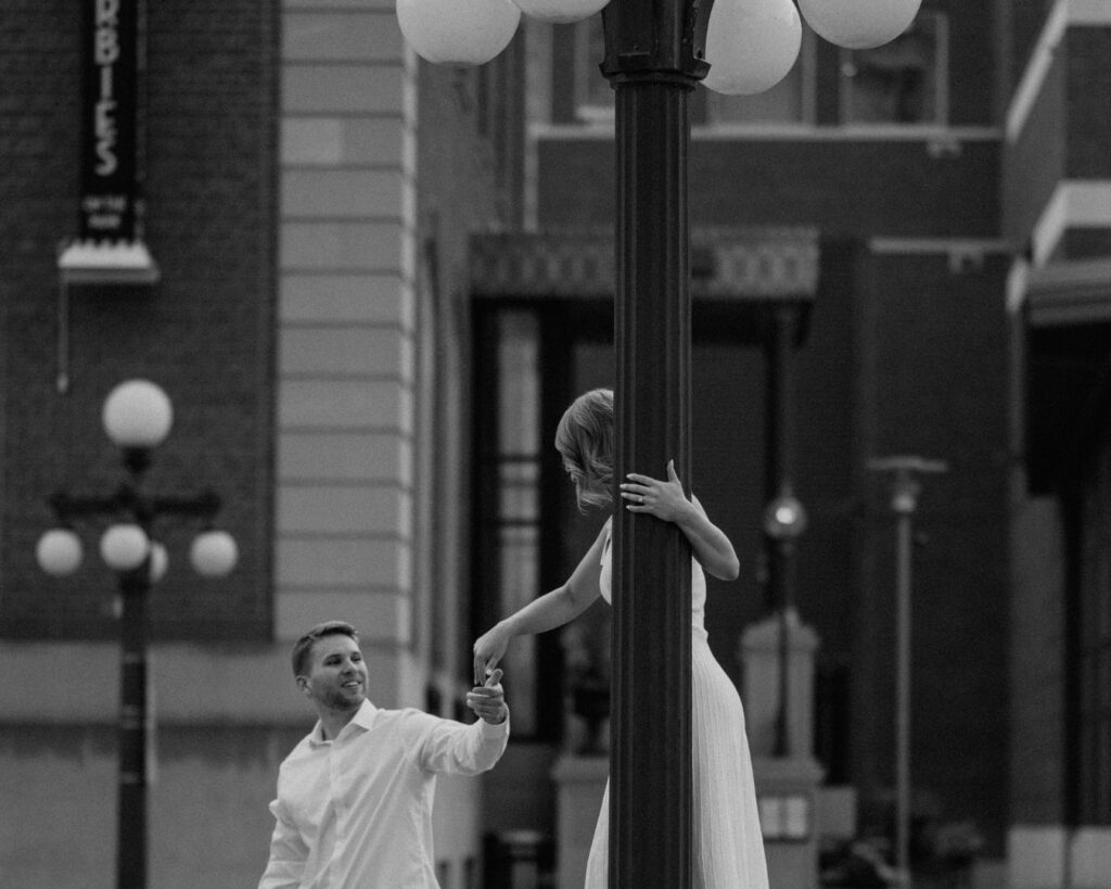 A man holding his fiance's hand as she spins around a vintage styled light pole. 