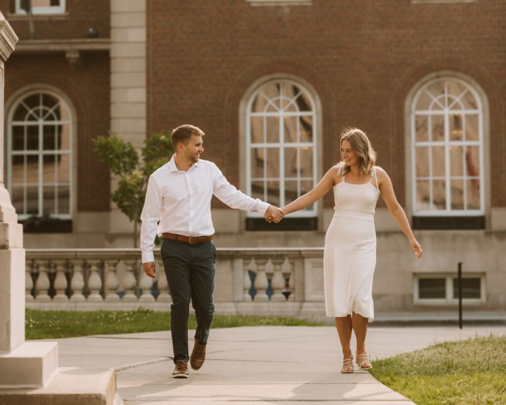 A couple walking hand in hand, grinning at each other in the city. 