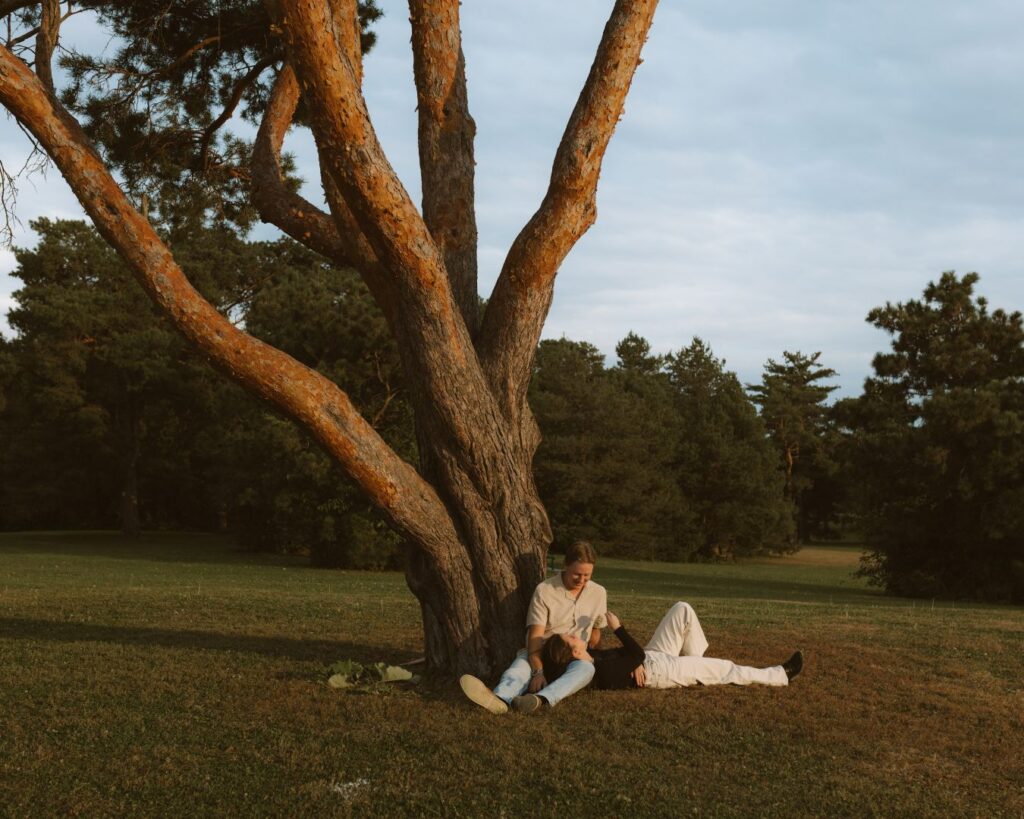 Romantic scene of a couple- he is sitting against a pine tree while she lays her head in his lap as they talk and laugh. 