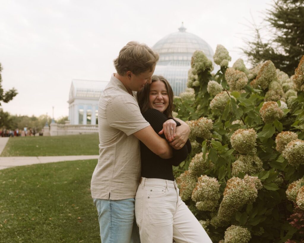 A man hugging his fiance from behind as they giggle at each other in front of the Como Zoo Conservatory. 